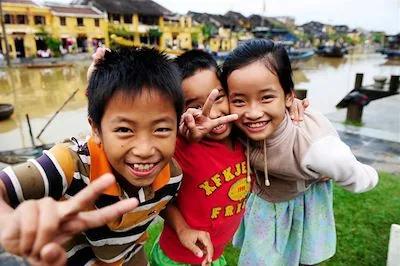 Trois enfants souriants font des signes devant un canal bordé de maisons anciennes